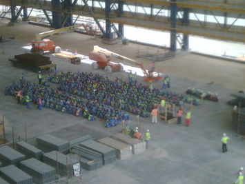 A large group of construction workers in high-visibility jackets and hard hats gathered sitting on the floor
