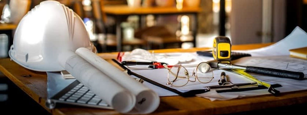 A cluttered construction desk with a hard hat, rolled blueprints, glasses, a tape measure, and a pen