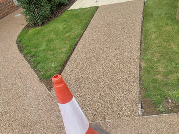 A newly laid resin-bound gravel path curving around a green lawn in a garden, with an orange traffic cone in the foreground