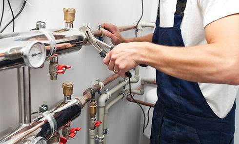 A plumber in blue overalls uses a wrench to adjust metal pipes with valves and a gauge