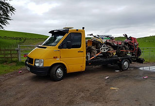 A yellow tow truck is parked on a rural road, carrying two ATVs