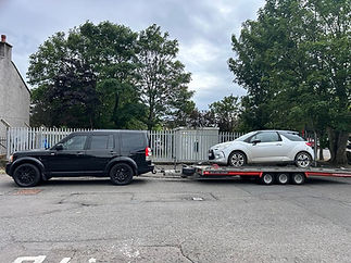 A black SUV tows a silver compact car on a red trailer in an urban area