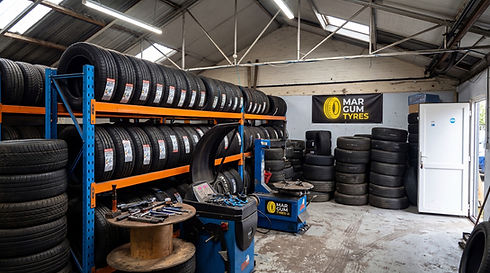 A tidy auto shop with stacked tires on shelves, a tire-changing machine, and tools on a wooden spool table