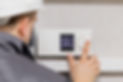 A person wearing a hard hat and jacket adjusts a digital thermostat on the wall