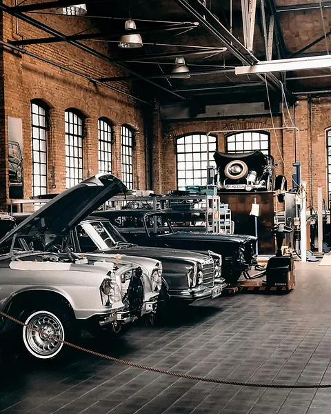 Several classic cars parked inside a spacious, brick-walled workshop, with the front vehicle showing its bonnet propped open for restoration