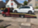 A white car is loaded on a red tow truck marked "24 HR Recovery" on a sunny day.