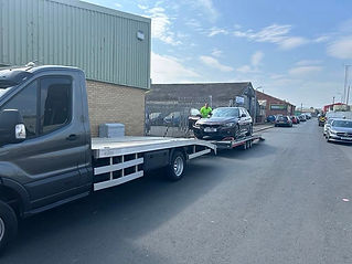 A tow truck with a flatbed carrying a black car is parked on an industrial street