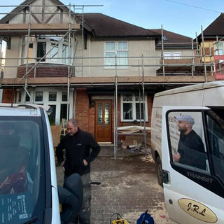 Two men stand beside white vans in front of a two-story house under renovation