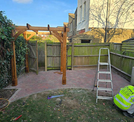 Wooden pergola on a brick patio next to a wooden fence. A worker in a yellow vest is crouching beside a ladder