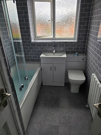 A compact bathroom featuring grey metro tiles, a bathtub with a glass screen, a white vanity unit sink, and a matching back-to-wall toilet
