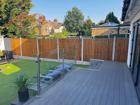 A raised grey composite decking area with a glass balustrade overlooking a lawned back garden