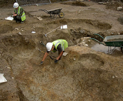 Two archaeologists in high-visibility vests and helmets work at an excavation site
