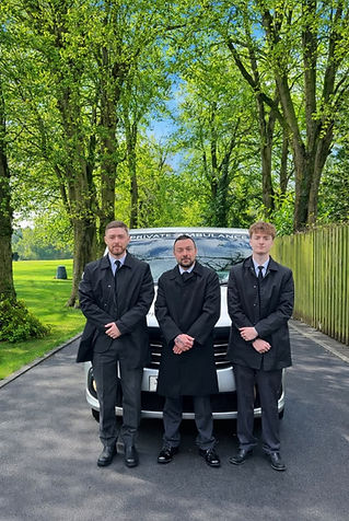 Three men in black suits and coats stand in front of a white van