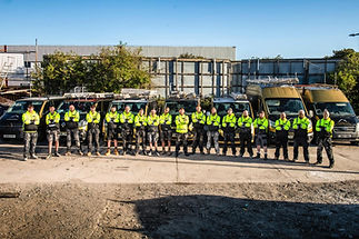 A group of 16 workers in matching high-visibility yellow jackets stand in front of work vans