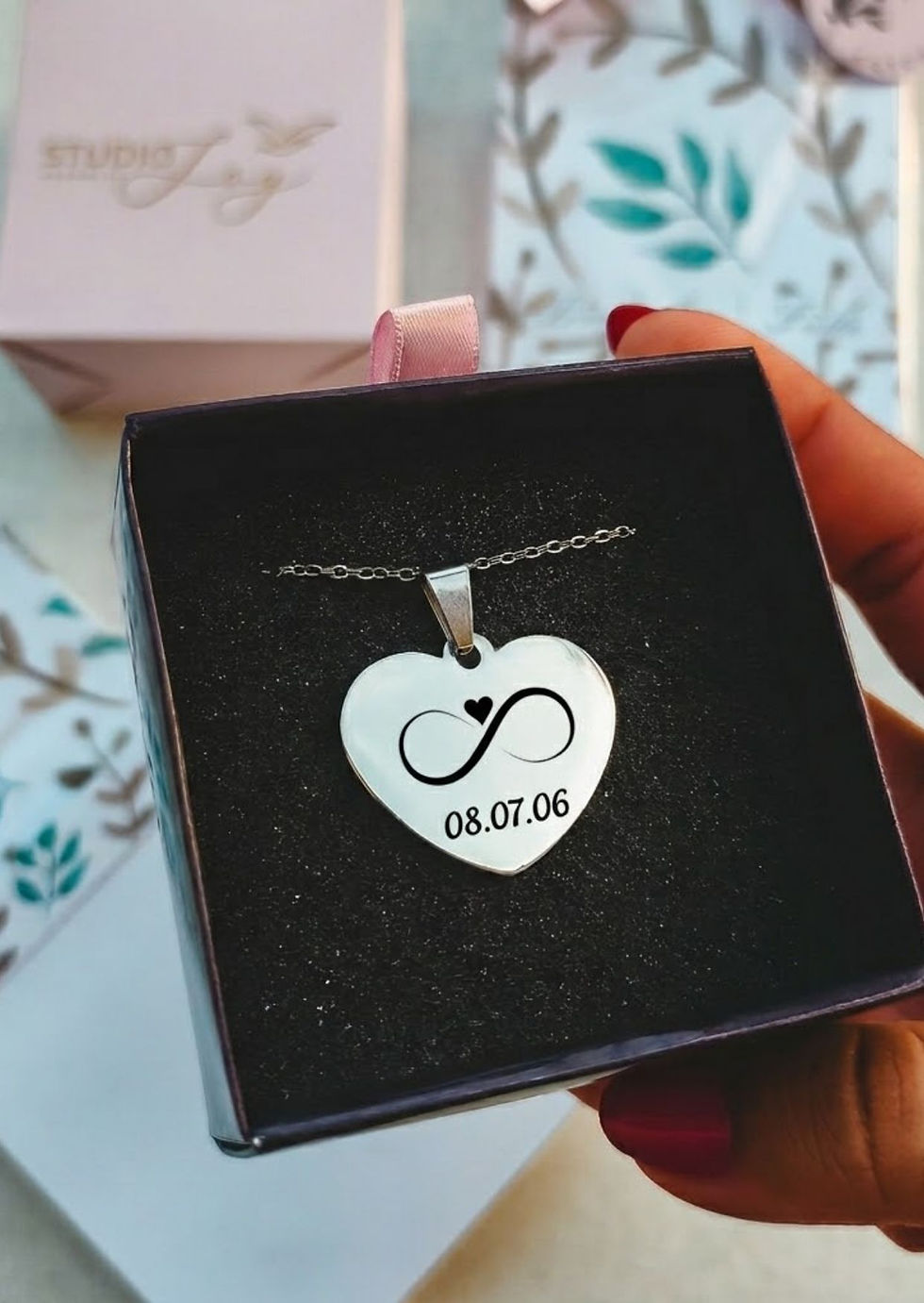 Mão segurando colar de aço inox com pingente de coração gravado com desenho infinito e data do casamento. Cordão Elo Portugue