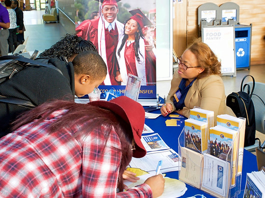 Three college-aged Black students (one with a pony tail, one in a black sweater and black back pack, and one in a red plaid shirt and red floppy hat, learning over a table filling out forms at an information table while a middle-aged Black femme or woman with glasses and light wavy hair watches.