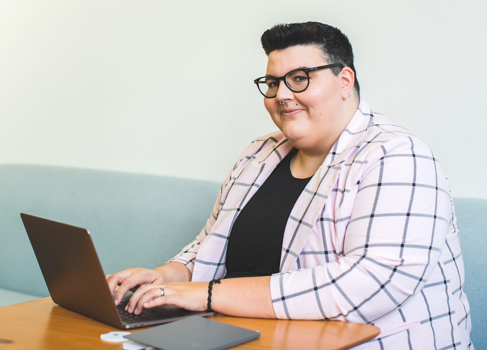 Photo of a light skinned non-binary person with short dark hair and thick glasses, smiling warmly while sitting at a laptop. They are wearing a striped pink shirt over a black t-shirt.