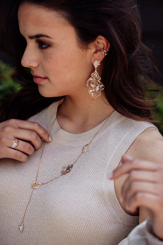 Young woman wearing matching 18k gold oak leaf necklace and earrings with pavé-set white diamonds – elegant nature-inspired.