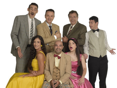 Seven people pose against a white backdrop, wearing vintage attire. Five men in suits and two girls in dresses; one in yellow and one in pink. Bright expressions.