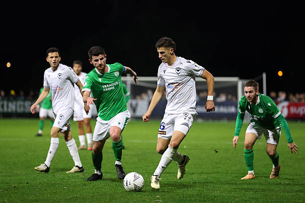 Soccer match at night; player in white dribbles ball while defended by players in green.