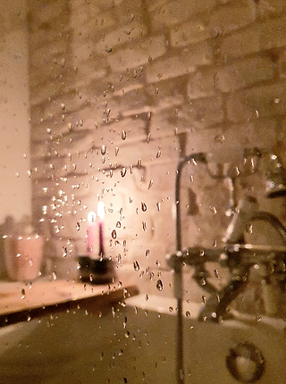 A close-up of a steamy bathroom mirror. Water droplets cling to the glass, obscuring the view of a bathtub and faucet. Two lit candles reflect in the mirror, creating a cosy atmosphere.