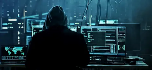 Silhouette of a person in a hoodie working in a dark room, backlit by the blue glow of multiple computer monitors displaying lines of code, creating an atmosphere of focused cybersecurity services, operations or hacking in an urban setting.