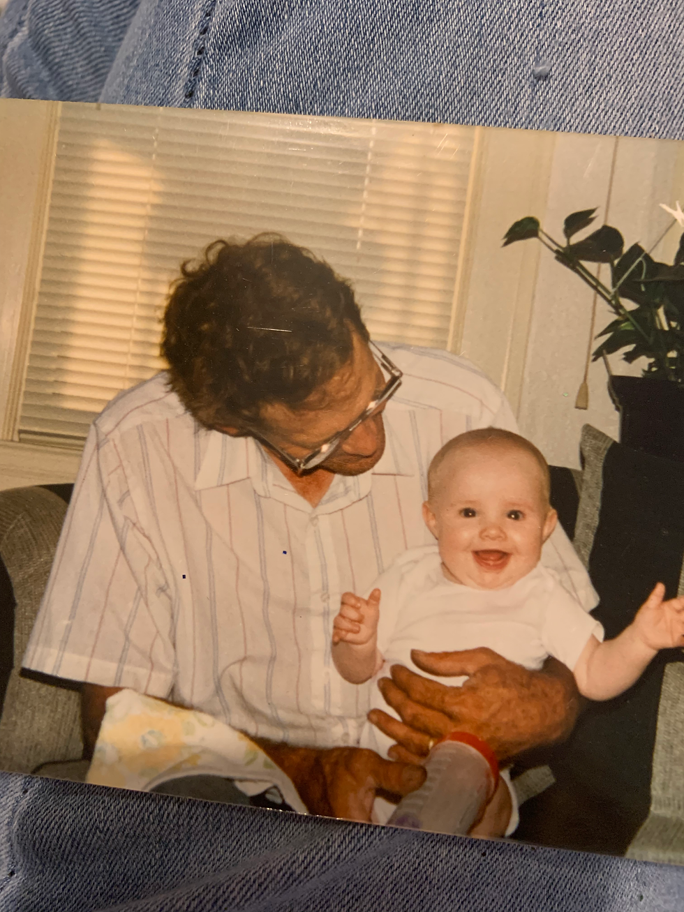 A man wearing glasses and a white and gray striped shirt is looking down at a baby sitting on his lap, holding a bottle to feed her. The baby is looking directly at the camera and has a big smile on her face. The baby is Emilie, and the man is her dad.
