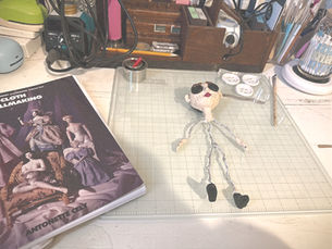 Work-in-progress handmade art doll on Emilie James's craft desk beside a cloth dollmaking book and art supplies.