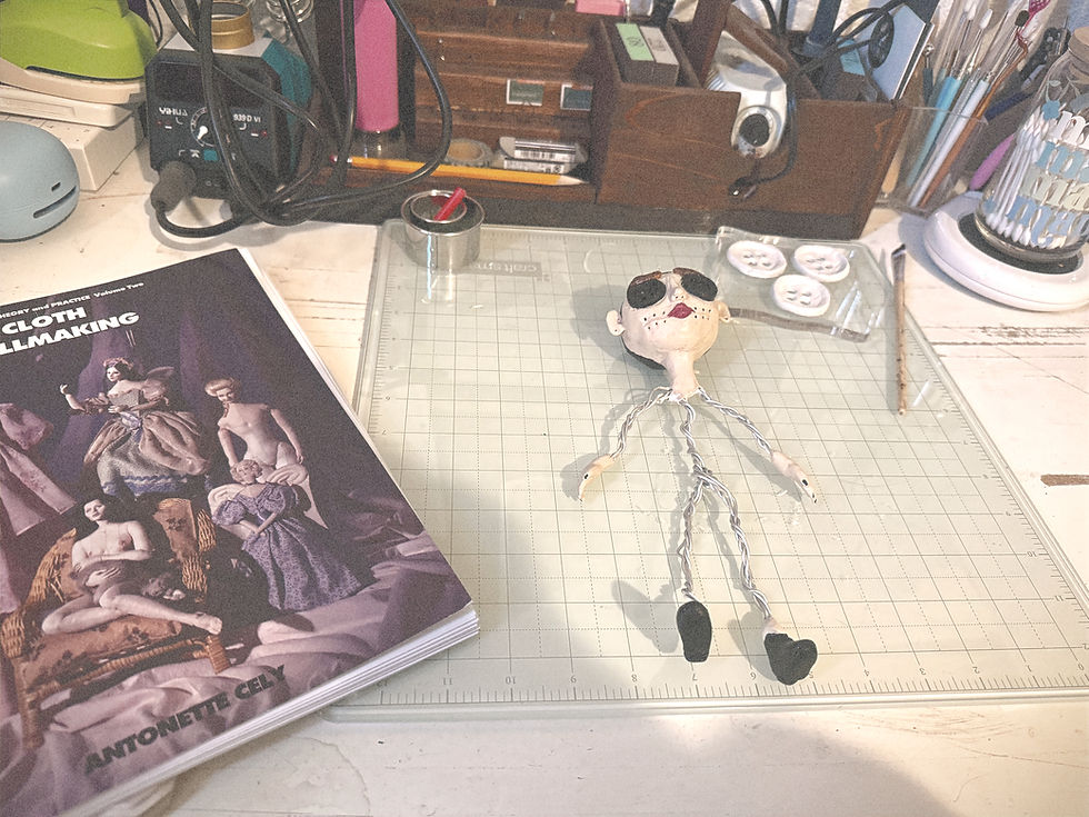 Work-in-progress handmade art doll on Emilie James's craft desk beside a cloth dollmaking book and art supplies.