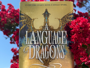 Emilie is holding up a copy of A Language of Dragons by S.F. Williamson against a tree with red flower petals and a blue sky dotted with clouds in the background. The cover is gold with a gold and blue dragon, wings outstretched from side to side.