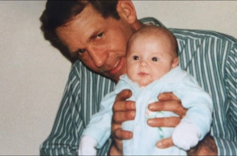 The year is 1996. A man with a blue and white striped shirt is holding a newborn infant dressed in blue. The man is holding the baby up like, "Look at what I got!" and he has a little smile. The baby is Emilie, and the man is her father.