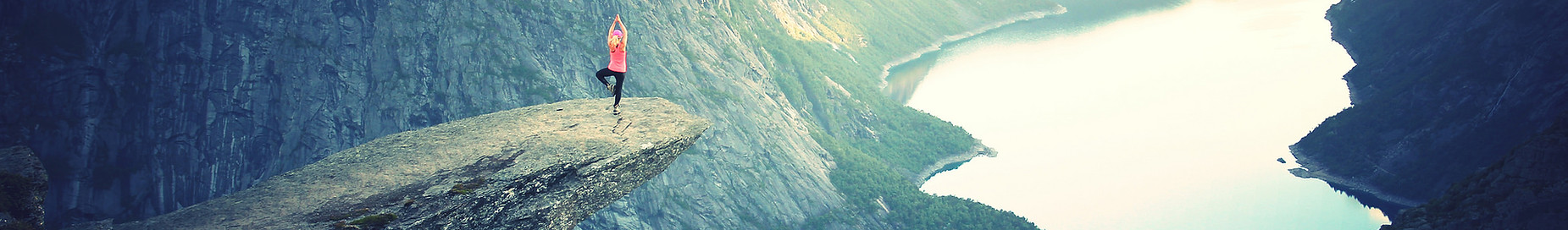 Jeune femme faisant du yoga sur un rocher au dessus du vide. Une vue imprenable sur un lac situé au bord d'une montagne est présente.