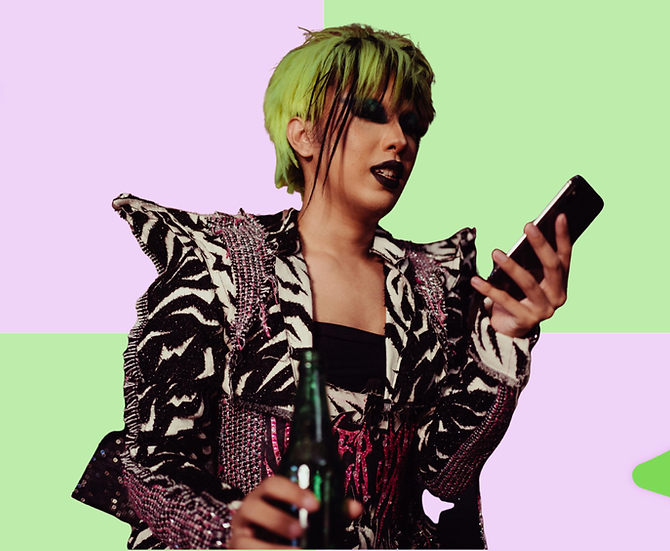 POC queer individual with outlandish jacket with shoulder pads and dark makeup including black lip and smoky eye, along with a pixie haircut dyed green and black. They are looking down at their phone.