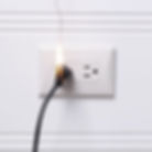 Photo of an electrical wall outlet with an electric cord plugged in. There is a flame rising out of the plug.