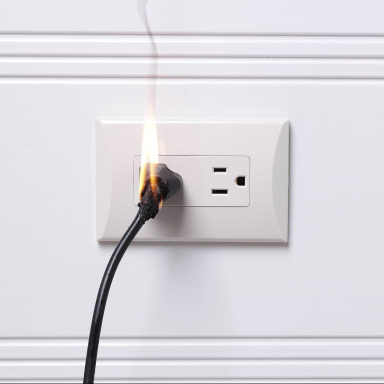 Photo of an electrical wall outlet with an electric cord plugged There is a flame rising out of the plug.