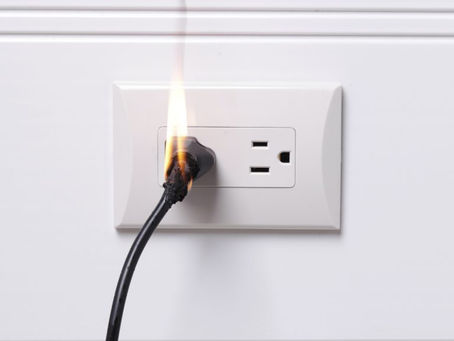 Photo of an electrical wall outlet with an electric cord plugged in. There is a flame rising out of the plug.