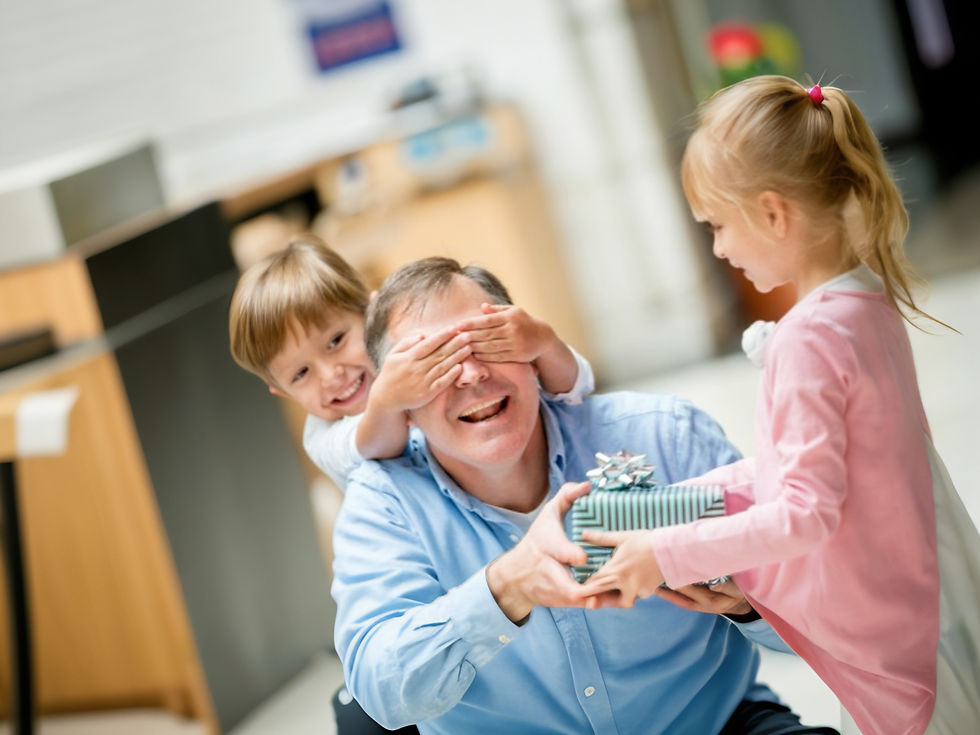 A young girl hands a present to an older man, while a young boy covers the man's eyes with his hands.