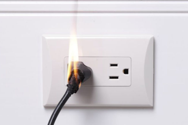 Photo of an electrical wall outlet with an electric cord plugged in. There is a flame rising out of the plug.