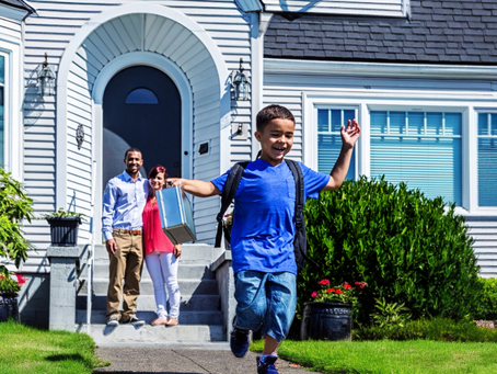 A man and woman watch is their child skips down their font walk. The child is wearing a backpack.