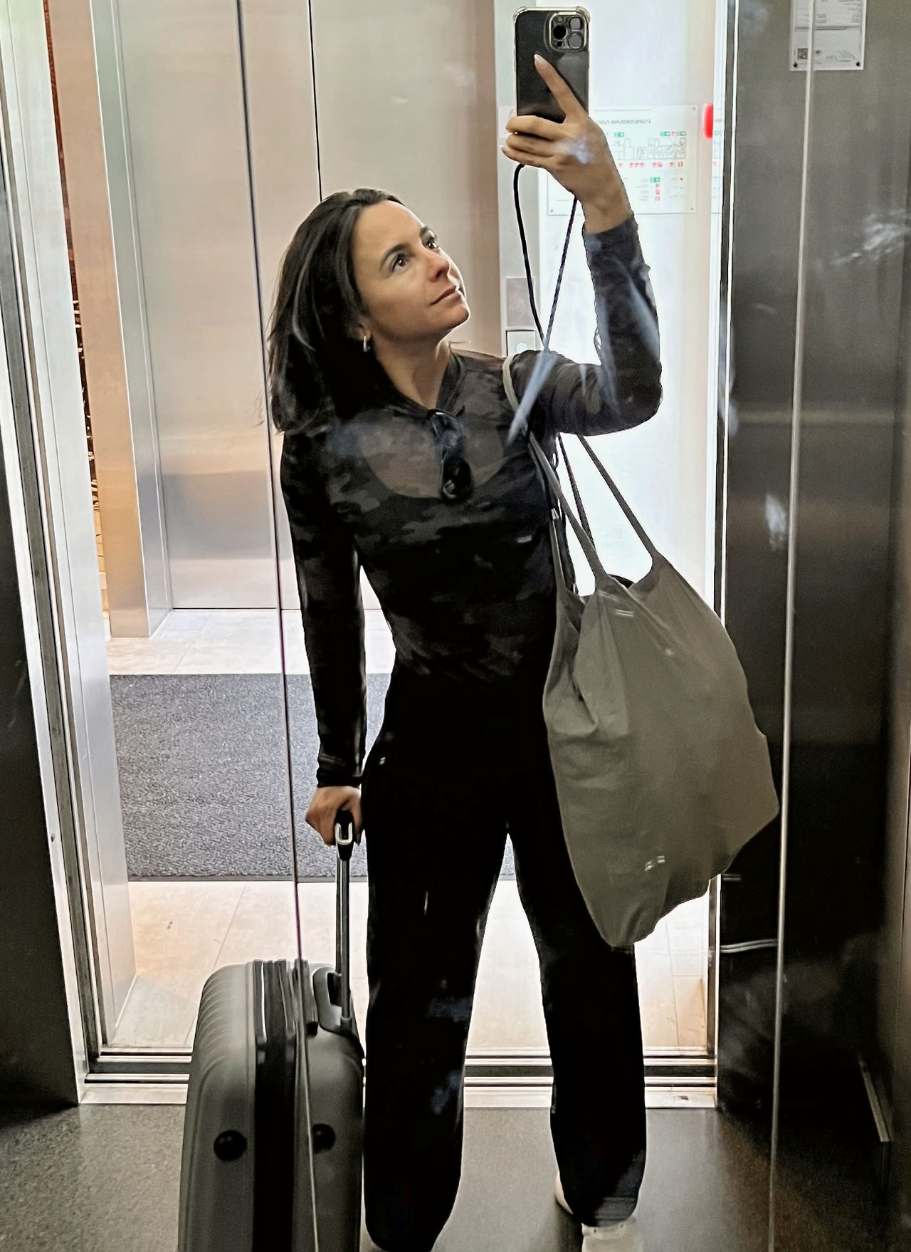 Foto no espelho de uma mulher vestida de preto com uma mala de rodinhas cinza e uma sacola bege.