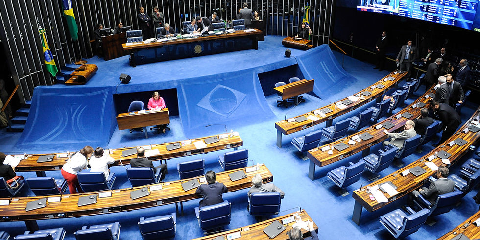 A imagem mostra o plenário do Senado Federal do Brasil, com seu característico carpete azul e assentos em disposição semicircular. No centro, a mesa diretora é ocupada por senadores, enquanto outros parlamentares se posicionam em suas respectivas cadeiras. A bandeira do Brasil está visível no canto superior esquerdo, e o brasão da República aparece gravado ao fundo, na parede de mármore. O ambiente é formal, com debates e discussões acontecendo, evidenciando o funcionamento do processo legislativo. O espaço transmite a atmosfera de decisões importantes sendo tomadas em nível nacional.