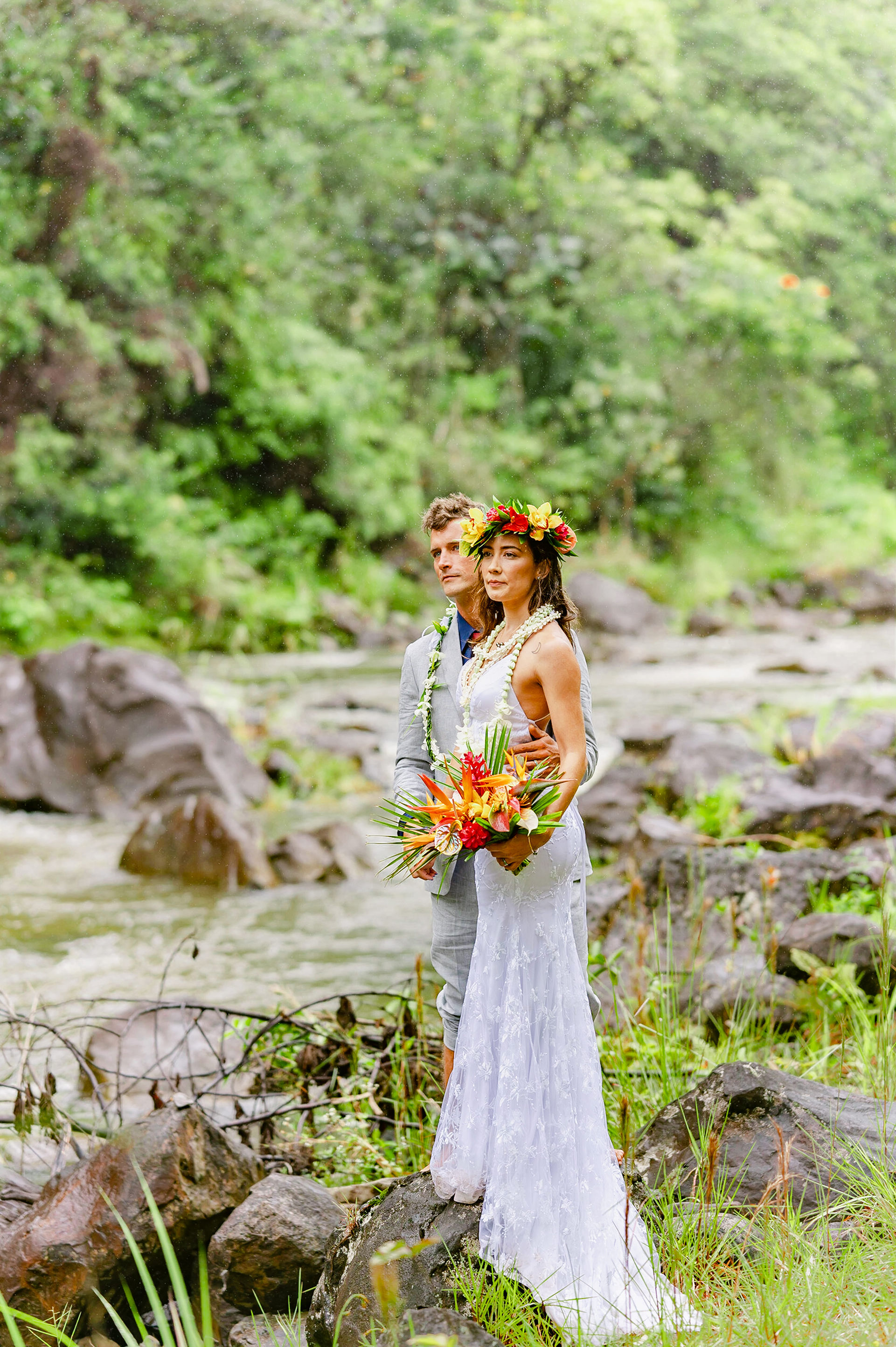 Couple embraces on rocky Kona shore at sunset, framed by ocean glow and lace wedding dress.