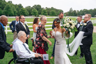 A bride holds a bouquet of flowers while smiling down at a guest. There is guests gathered around with a white fence in the background.