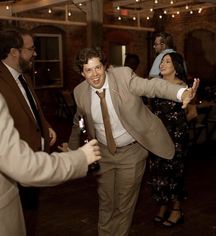 A man in a kakhi suit smiles at the camera. He is slightly bent over, holding his arm out. There is guests behind him.