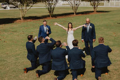A bride poses in front of a group of groomsmen. She holds her arms out in the center while they face her.