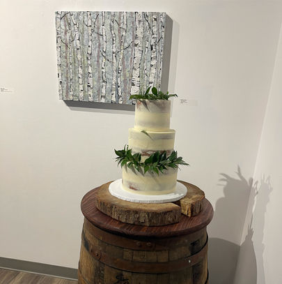 A white three tier wedding cake with greenery on top of two of the tiers, on a wood slab cake stand and wooden whiskey barrel in front of a painting of grey birch trees 