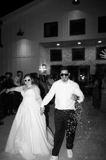 A bride and groom pose together in front of a building. The bride wears a white strapless gown and the groom wears a white suit coat. There is guests cheering around them.