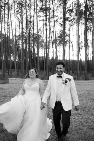 Black and white photo of a bride and groom holding hands walking together. They are outside with a forest behind them. The bride wears a white strapless gown. The groom wears a white jacket with a black bowtie.