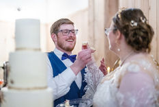 A bride and groom are holding cake in each of their hands. They both are feeding the cake to each other. A three tier white wedding cake is pictured beside them with gold detailing. 