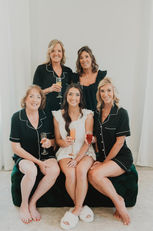 A bride and four bridesmaids sit on a couch wearing pajamas. The bride wears white and the rest of the women wear black sets.